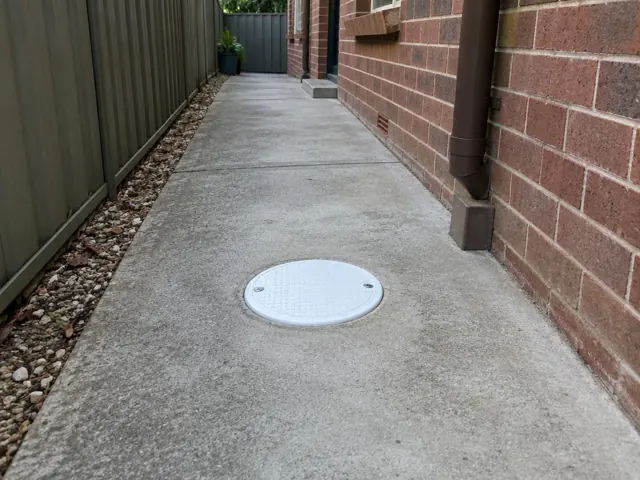 Concrete side path beside brick wall with a capped drain inspection opening in Auburn