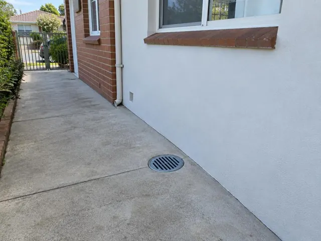 Outdoor concrete path with a round drain grate beside brick and painted walls in Berala